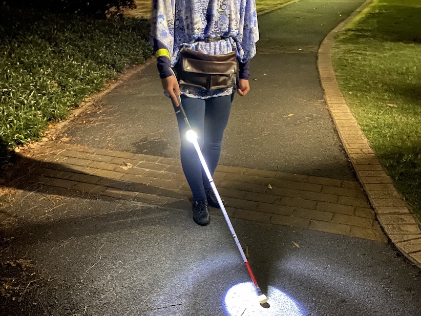 Woman holding a long cane with a light walking along a path in the evening