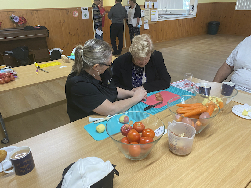 Lisa sitting with Dorothy at a table with vegetables and chopping boards