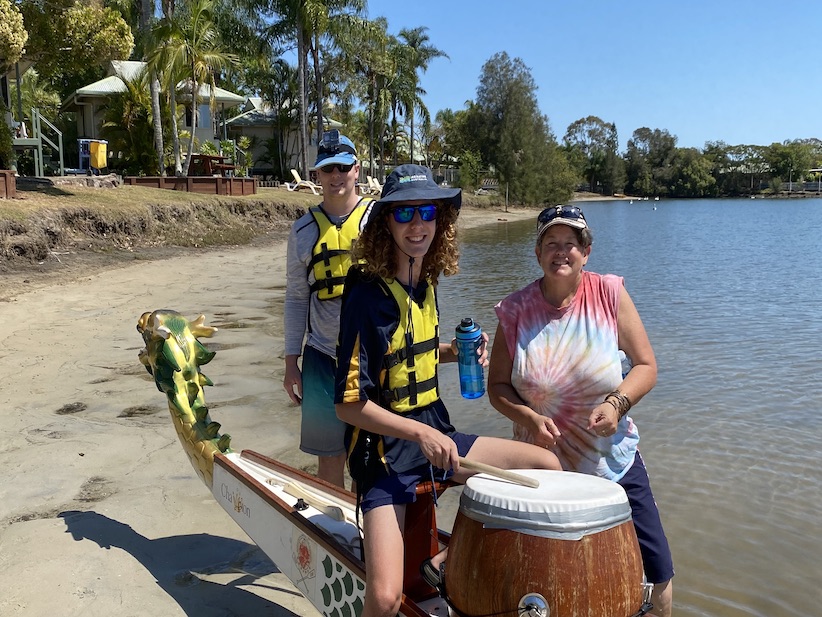 Group of three people at the rivers edge with a dragon boat and conga drum