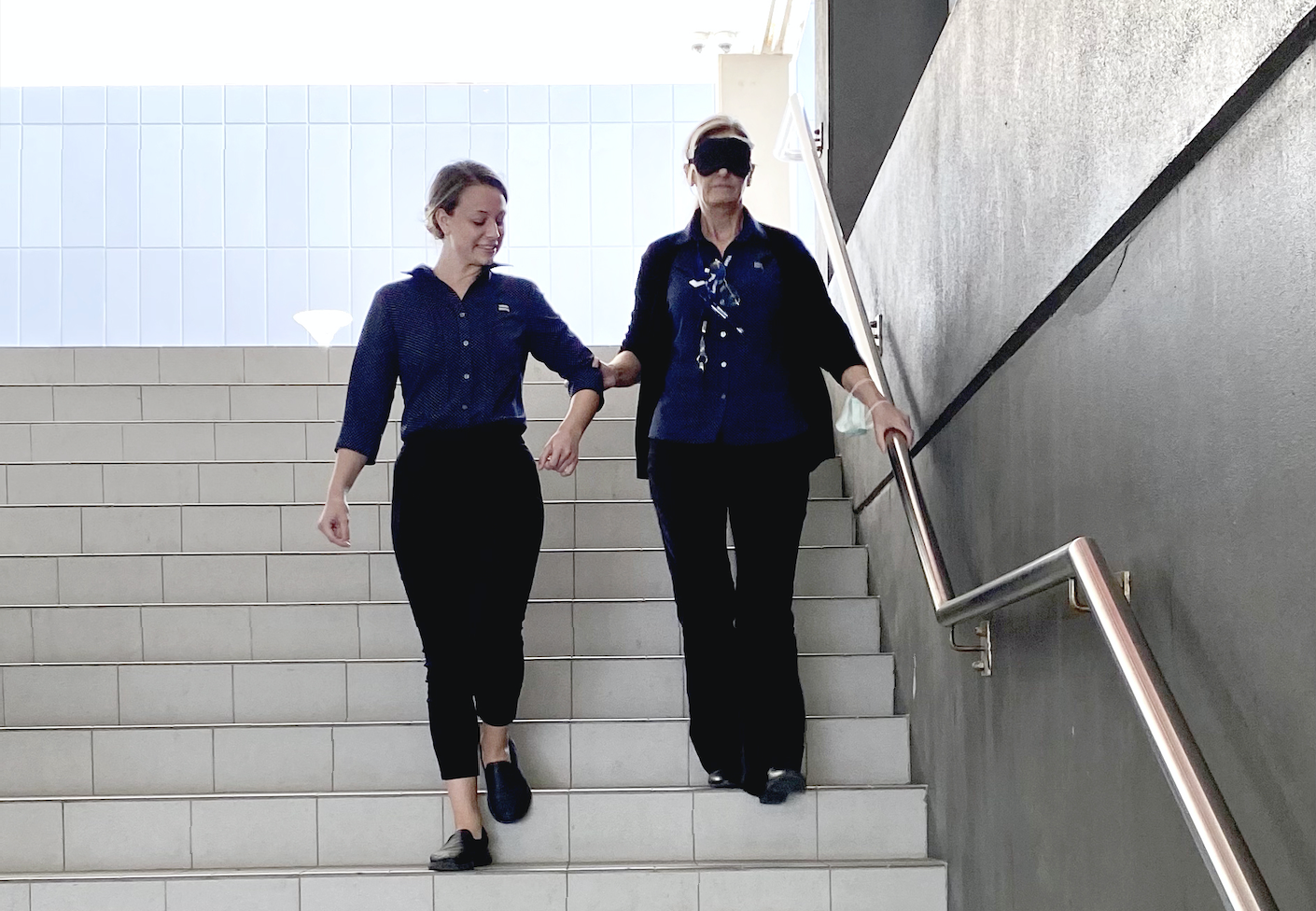 Photo of two people walking downstairs with one blind folded and the other guiding her.