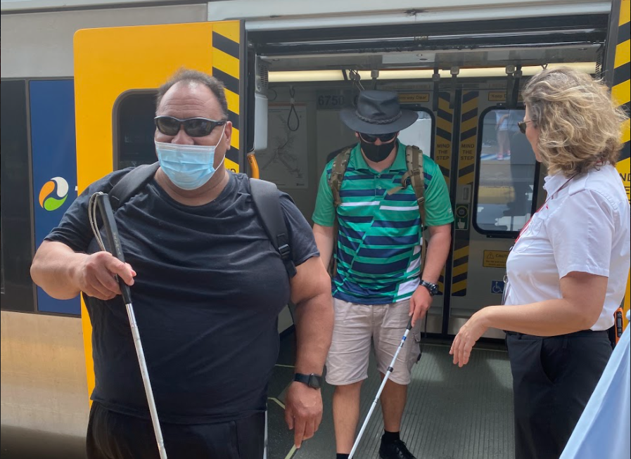 Two travellers with long canes get off the train with door held open by Qld Rail Staff